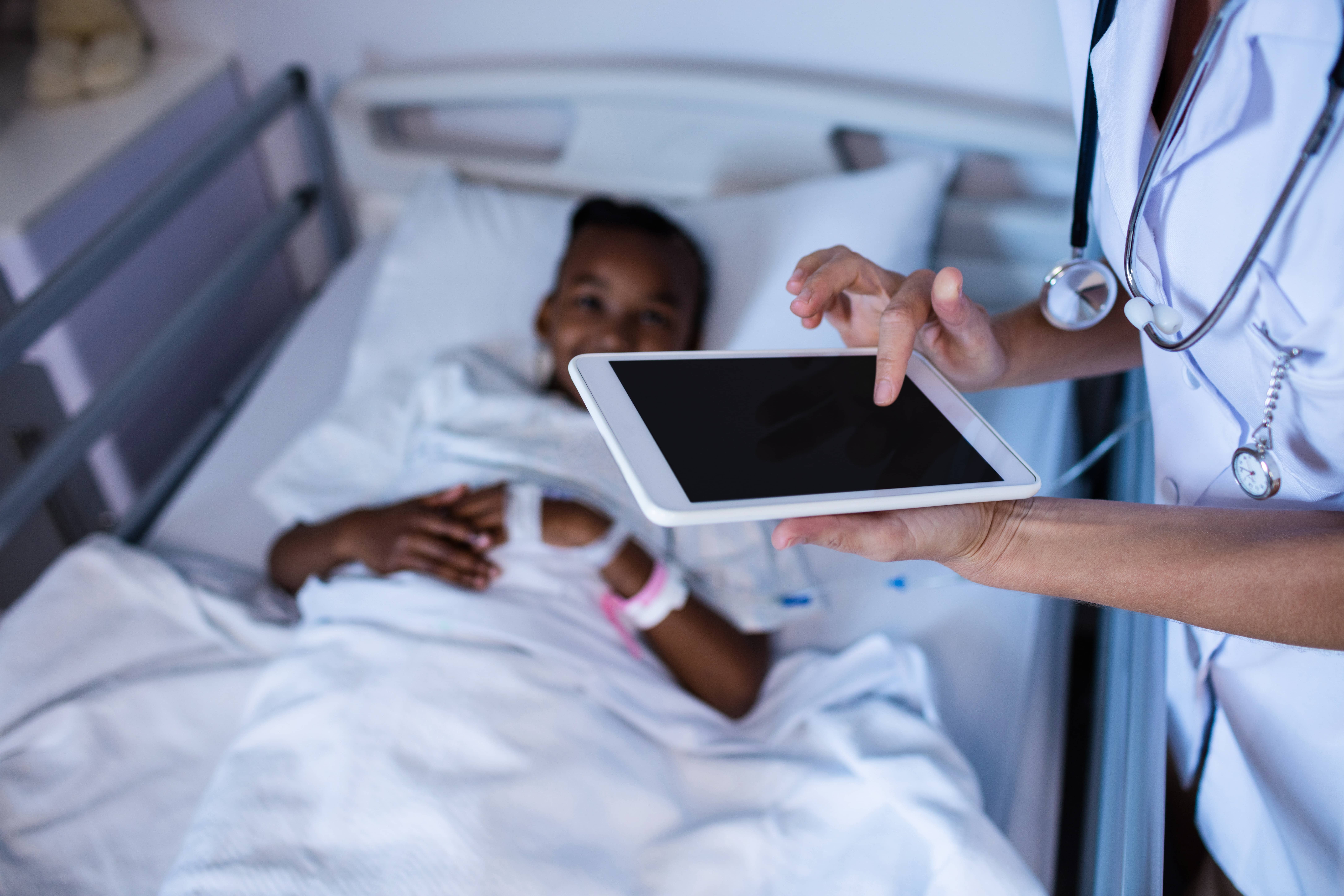 Um quarto de hospital, com uma criança negra feliz deitada em uma maca. Ao seu lado, mas sem aparecer o rosto, há uma enfermeira com um tablet na mão clicando na tela do disposito.